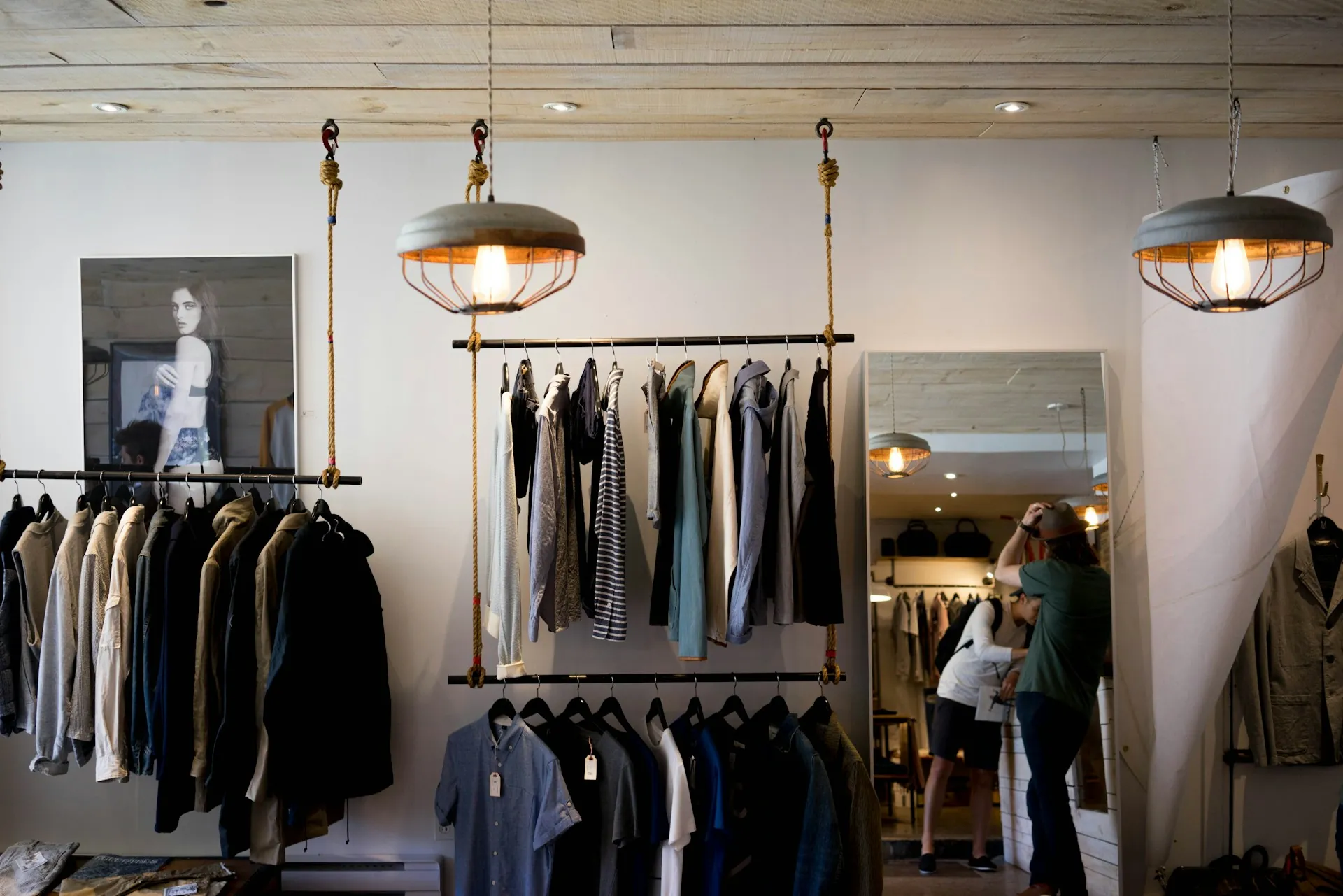 Independent clothing boutique interior with hanging racks, industrial pendant lights, and customers browsing